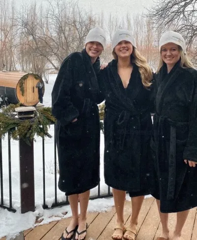 Three women in black robes and white hats stand on a snowy deck near a barrel sauna
