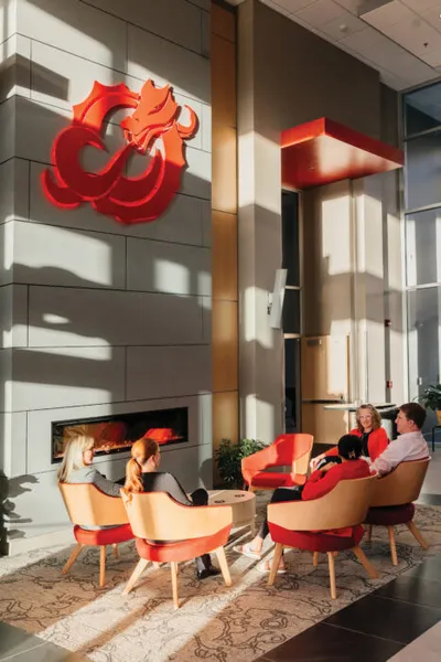 Four people seated in red and tan chairs around a low table in a sunlit lobby with a fireplace
