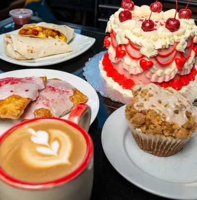 Latte with latte art beside a cherry-topped layer cake, glazed muffin, pastries, and a breakfast wrap