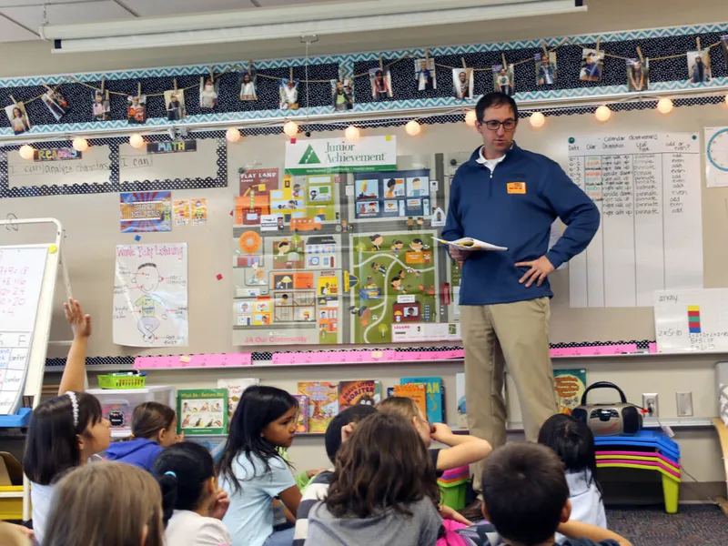 Man in blue sweater leads a Junior Achievement lesson for a group of young students in a classroom