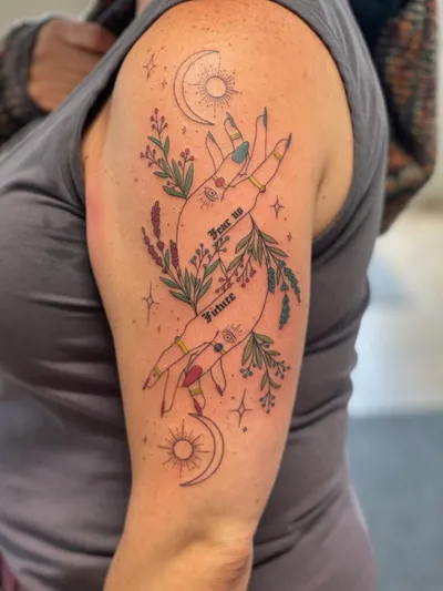 Colorful upper-arm tattoo of two hands with floral sprigs, moons, and a cursive banner