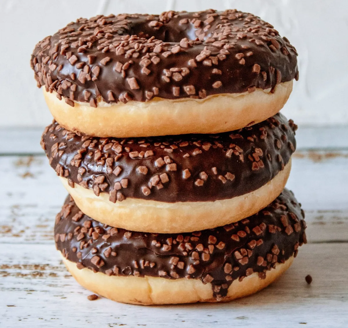 Stack of three chocolate-glazed donuts with chocolate sprinkles on a white wooden surface