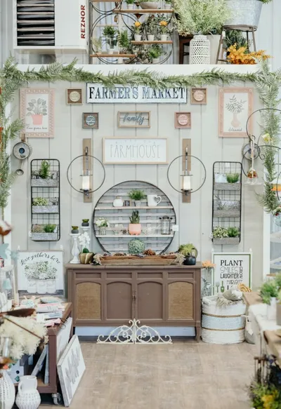 Farmhouse-style retail display with a 'Farmer's Market' sign, wooden cabinet, potted plants, and rustic wall décor