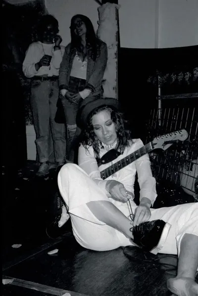 Woman in white outfit and hat sits on stage floor playing electric guitar while two people watch from behind