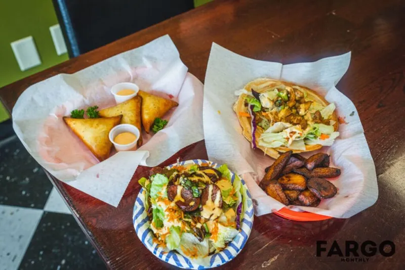 Fried samosas with dipping sauce, a loaded taco, a salad with sausage, and fried plantains on a wooden table