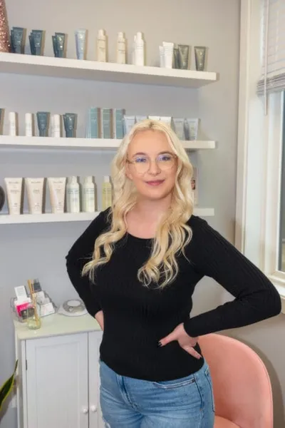 Blonde woman in black top and jeans poses in a skincare clinic with product shelves behind her