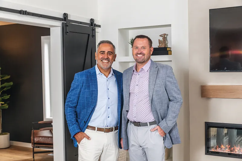 Two smiling men in sport coats standing together inside a modern home with a barn door and fireplace