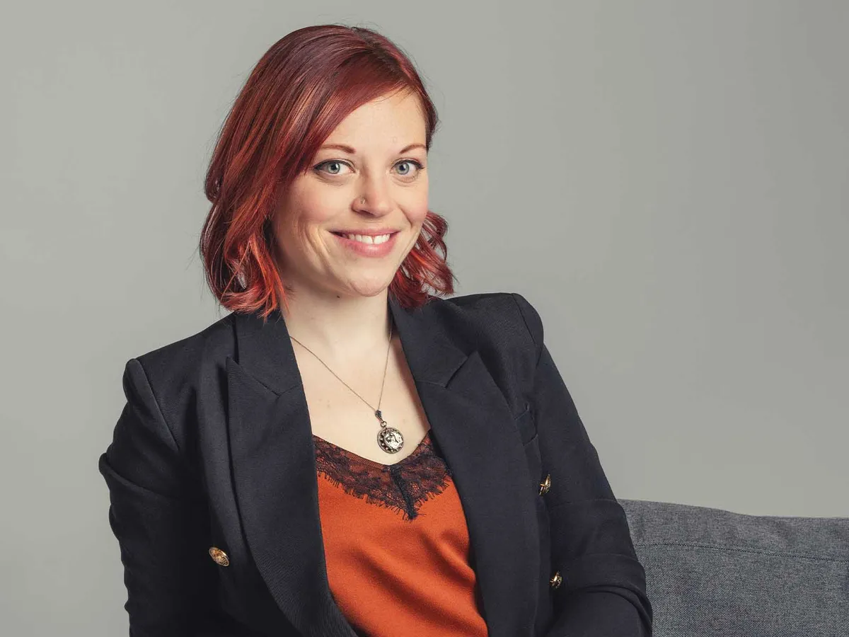 Smiling woman with red hair in a black blazer and rust lace-trim top seated on a grey sofa