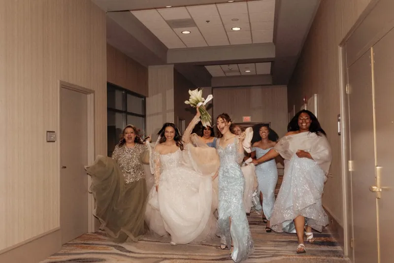 Bride in white gown raises bouquet while bridesmaids in blue and sage cheer in a hotel corridor