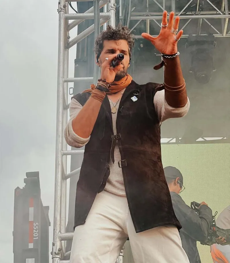 Male singer performs on outdoor stage, mic in hand, one arm raised, wearing a dark vest and orange scarf