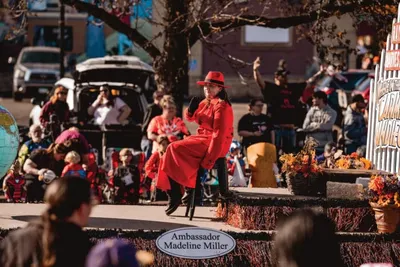 Woman in red dress and hat waves from a parade float labeled Ambassador Madeline Miller
