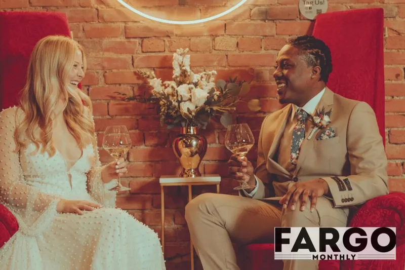 Bride and groom laughing and clinking wine glasses in red velvet chairs against a brick wall