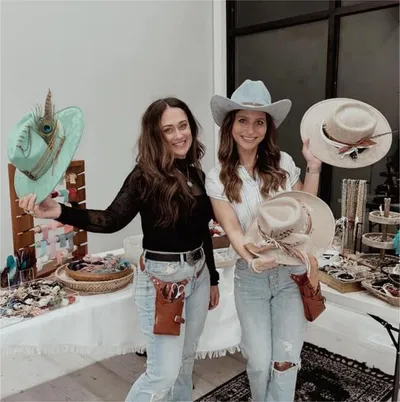 Two smiling women holding decorated cowboy hats near a table of accessories