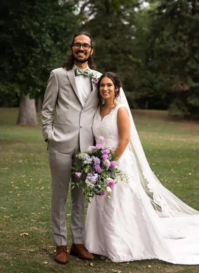 Bride in white gown holding purple bouquet stands with groom in light gray suit outdoors