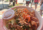 Bowl of noodle salad with colorful toppings and a dipping sauce at an outdoor market