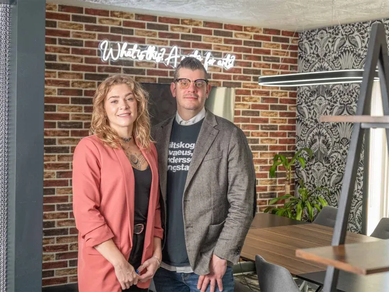 A woman in a pink blazer and a man in a gray blazer stand before an exposed brick wall with a neon sign