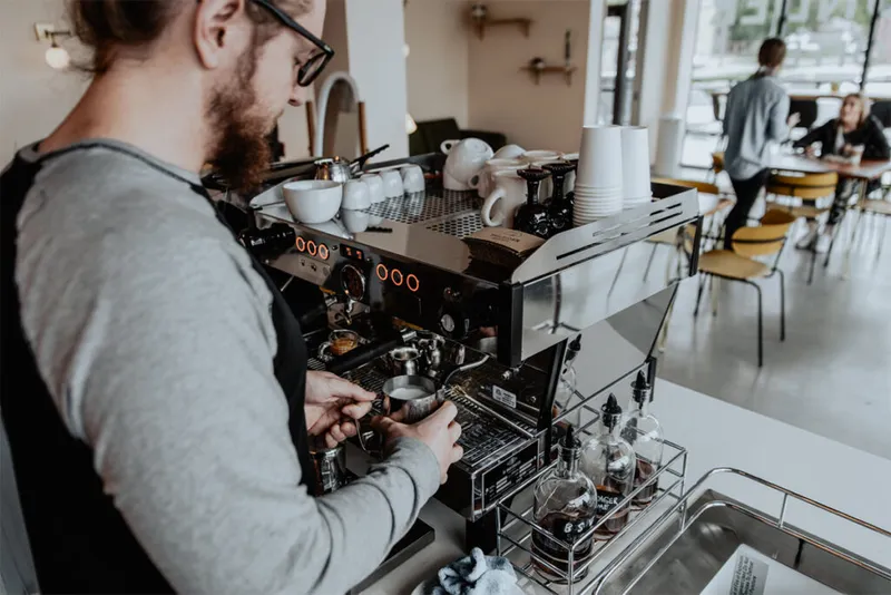 Bearded barista in apron steaming milk at a commercial espresso machine in a bright café
