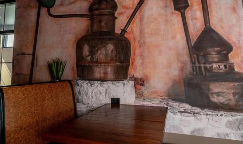 Restaurant booth with wood table beside a large mural of copper distillery stills