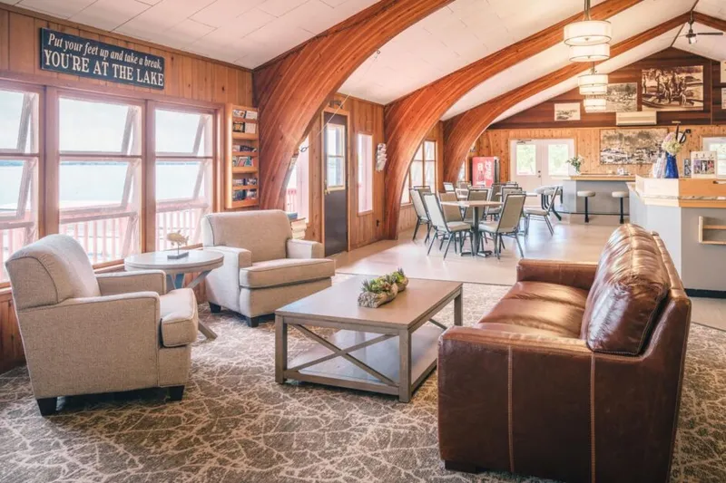 Rustic lodge lobby with leather sofa, beige armchairs, arched wood beams, and a 'You're At The Lake' sign