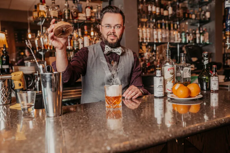 Bearded bartender in bow tie and vest holds a garnish over a smoking cocktail at a marble bar
