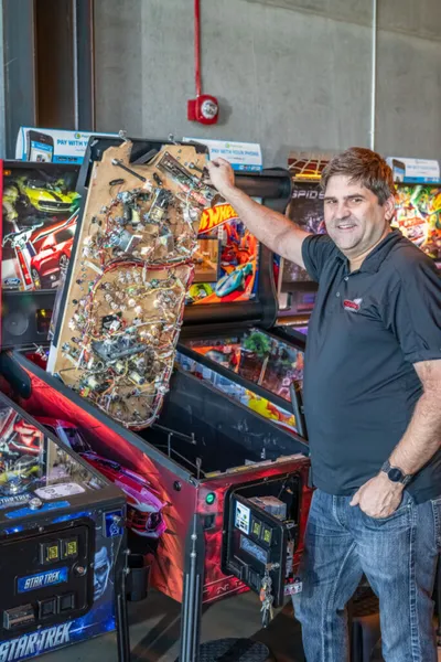 Man in black polo lifts the playfield of a pinball machine in an arcade