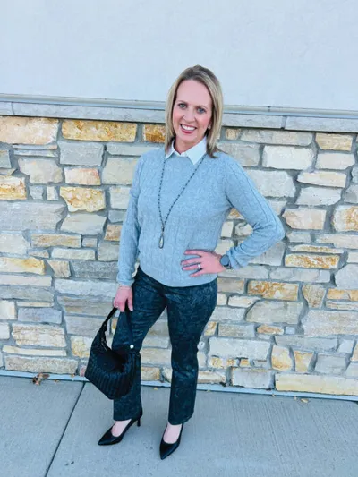 Blonde woman in gray cable-knit sweater, dark patterned pants, and black heels holding a black woven bag in front of a stone