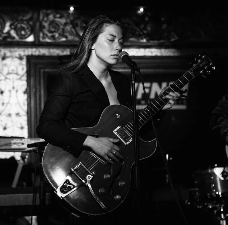 Woman with long hair playing a hollow-body electric guitar at a dimly lit venue