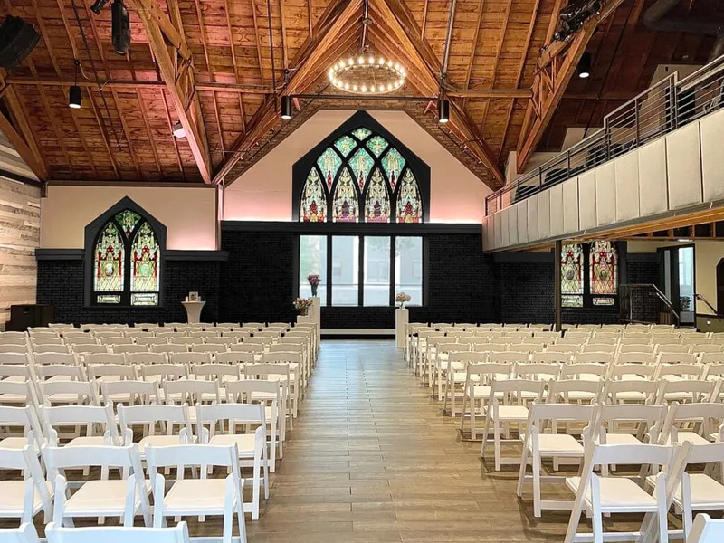 Rows of white folding chairs line a center aisle in a chapel-style venue with stained glass windows and exposed wood ceiling