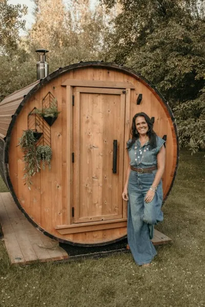 Woman in denim outfit smiling beside a round cedar barrel sauna outdoors