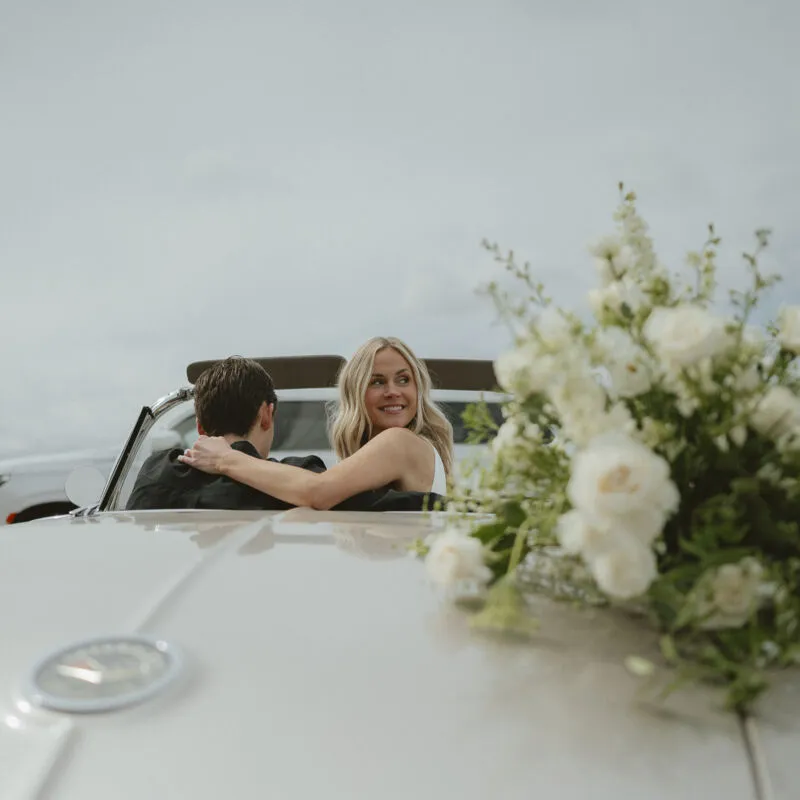 Smiling blonde woman embraces a man in a white vintage convertible, white floral arrangement in foreground