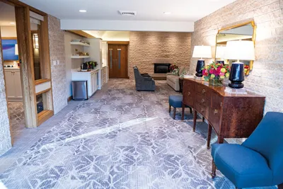 Spacious funeral home lounge with patterned carpet, stone fireplace, wood console, and blue chairs