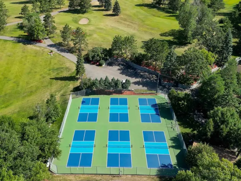 Aerial view of six blue pickleball courts on a green surface surrounded by trees and a golf course