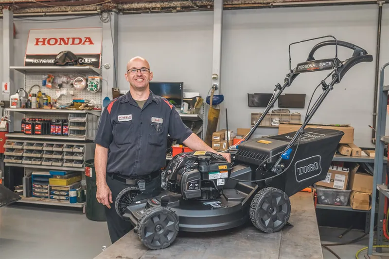 Smiling technworker in a shop uniform stands beside a Toro Personal Pace lawn mower in a repair workshop