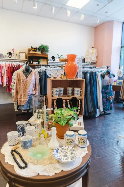 Boutique interior with clothing racks, mannequins, and a table displaying ceramic mugs, vases, and a potted plant