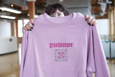 Person holding up a lavender sweatshirt printed with "grandmacore" and a quilt-style graphic