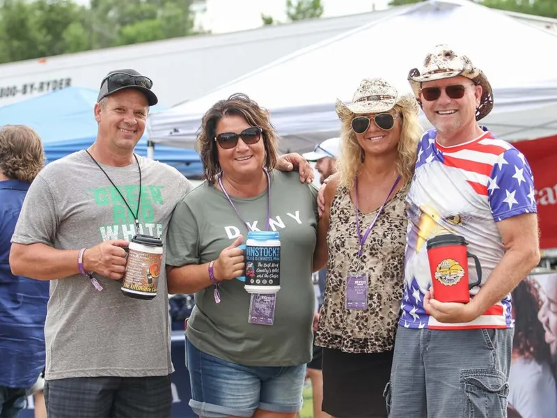 Four smiling festival-goers holding drinks, two wearing cowboy hats, one in an American flag shirt