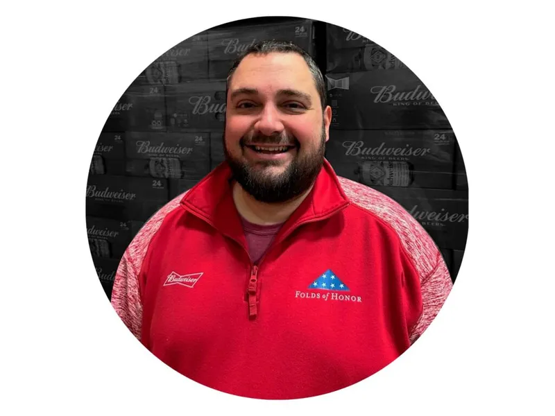 Smiling bearded man in a red Budweiser Folds of Honor quarter-zip, stacked Budweiser cases behind him