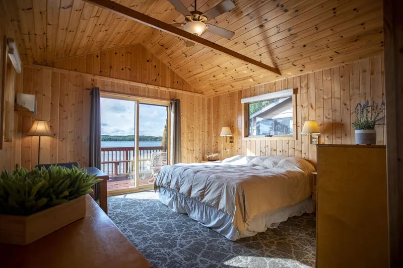 Pine-paneled cabin bedroom with vaulted ceiling, queen bed, and sliding doors opening to a lakeside deck