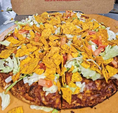 Taco pizza topped with tortilla chips, shredded lettuce, and diced tomatoes in a cardboard box