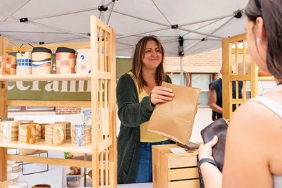 Smiling woman hands a paper bag to a customer at an outdoor ceramics market stall