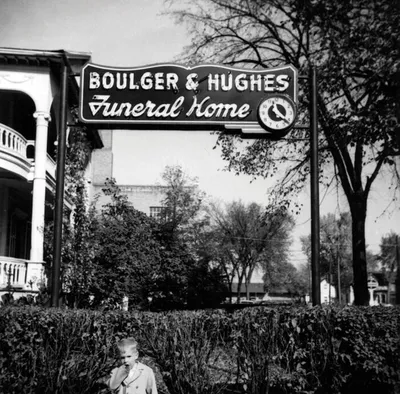Child stands beneath a vintage Boulger & Hughes Funeral Home sign in a black-and-white street scene