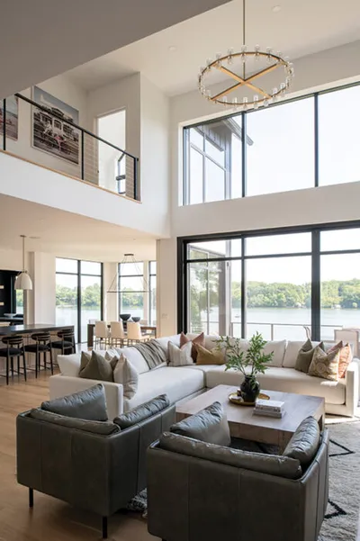 Airy double-height living room with white sectional, gray leather chairs, and floor-to-ceiling windows overlooking a lake