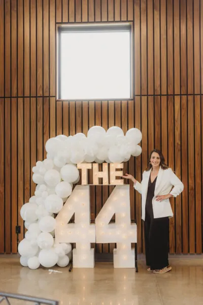 Woman in white blazer posing beside illuminated "The 44" marquee letters with white balloon arch