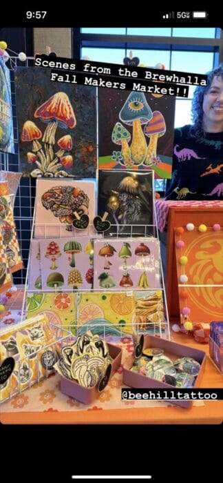 Artist smiling behind a market table displaying colorful mushroom-themed prints, stickers, and small goods
