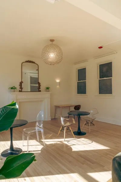 Sunlit co-working lounge with round bistro tables, clear acrylic chairs, and a globe pendant light above a white fireplace