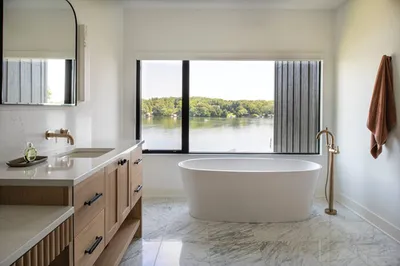 Freestanding white soaking tub before a large window overlooking a lake and trees