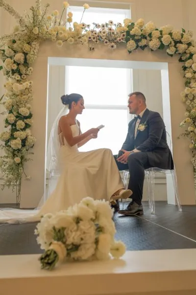 Bride reading vows to groom seated beneath a white floral arch during a wedding ceremony