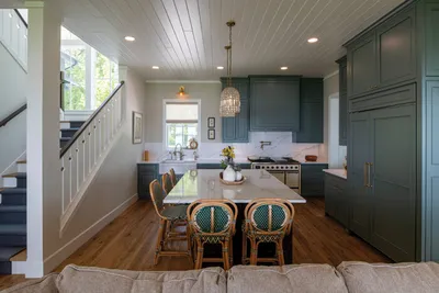 Open kitchen with dark green cabinetry, white island, rattan barstools, and wood floors beside a staircase