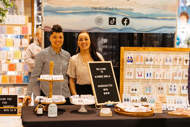 Two women smile behind a craft fair booth displaying handmade earrings and a Rubee Soul welcome sign