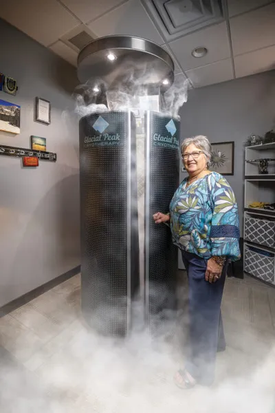Smiling older woman standing beside a cylindrical cryotherapy chamber emitting cold vapor mist